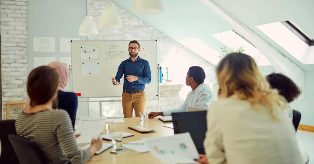 A Business Change expert stands in front of a whiteboard in a meeting to brief a team of people