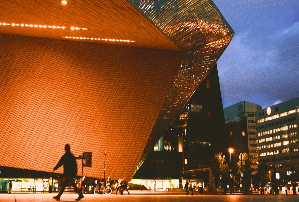 A modern building with orange walls and shiny trim, a dark sky and people walking on the streets below